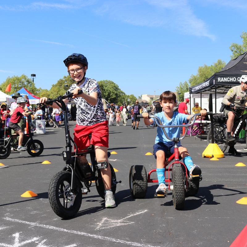 Children enjoying a biking activity at a busy outdoor event with crowds and tents in the background.