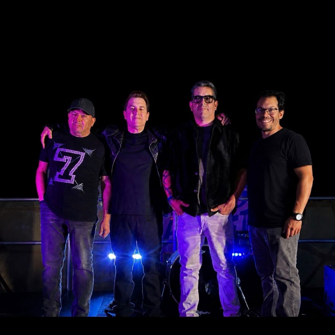  Four people standing side by side on a stage at night, illuminated by bright blue lights with musical equipment visible behind them.